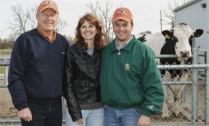 Charlie Will (à esquerda) com Brian e Wendy Fust, criadores de Fustead Emory BLITZ, na Select Sires. Atrás deles, o próprio Fustead Emory BLITZ se destaca como prova do olhar de Charlie para "diamantes brutos" — um touro que venderia mais de 1.52 milhão de unidades para a Select Sires, um recorde da empresa.